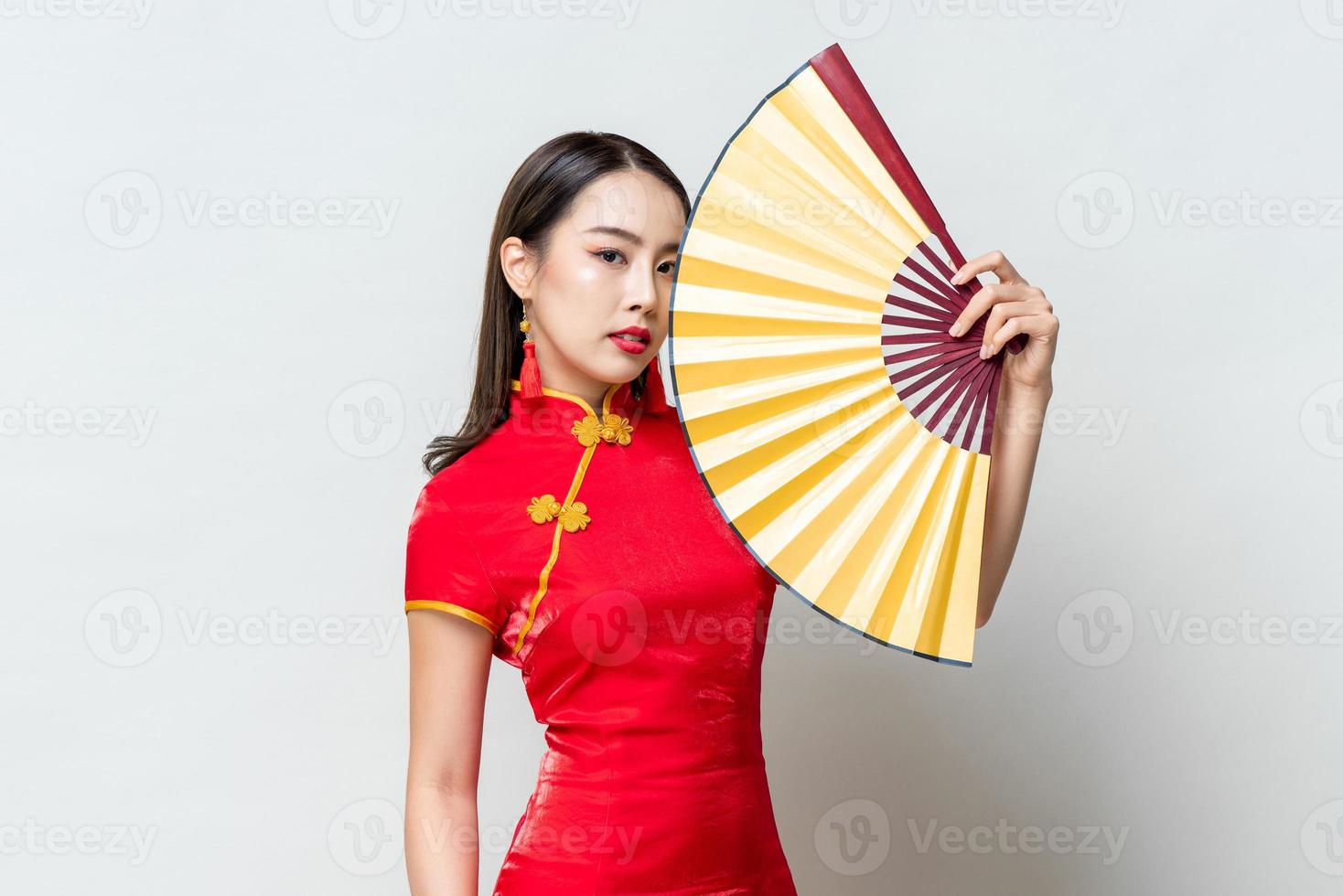 porträt der schönen asiatischen frau, die traditionelles rotes qipao-kleid trägt, das goldenen fan im isolierten hellgrauen hintergrund des studios für chinesisches neujahrskonzept hält foto