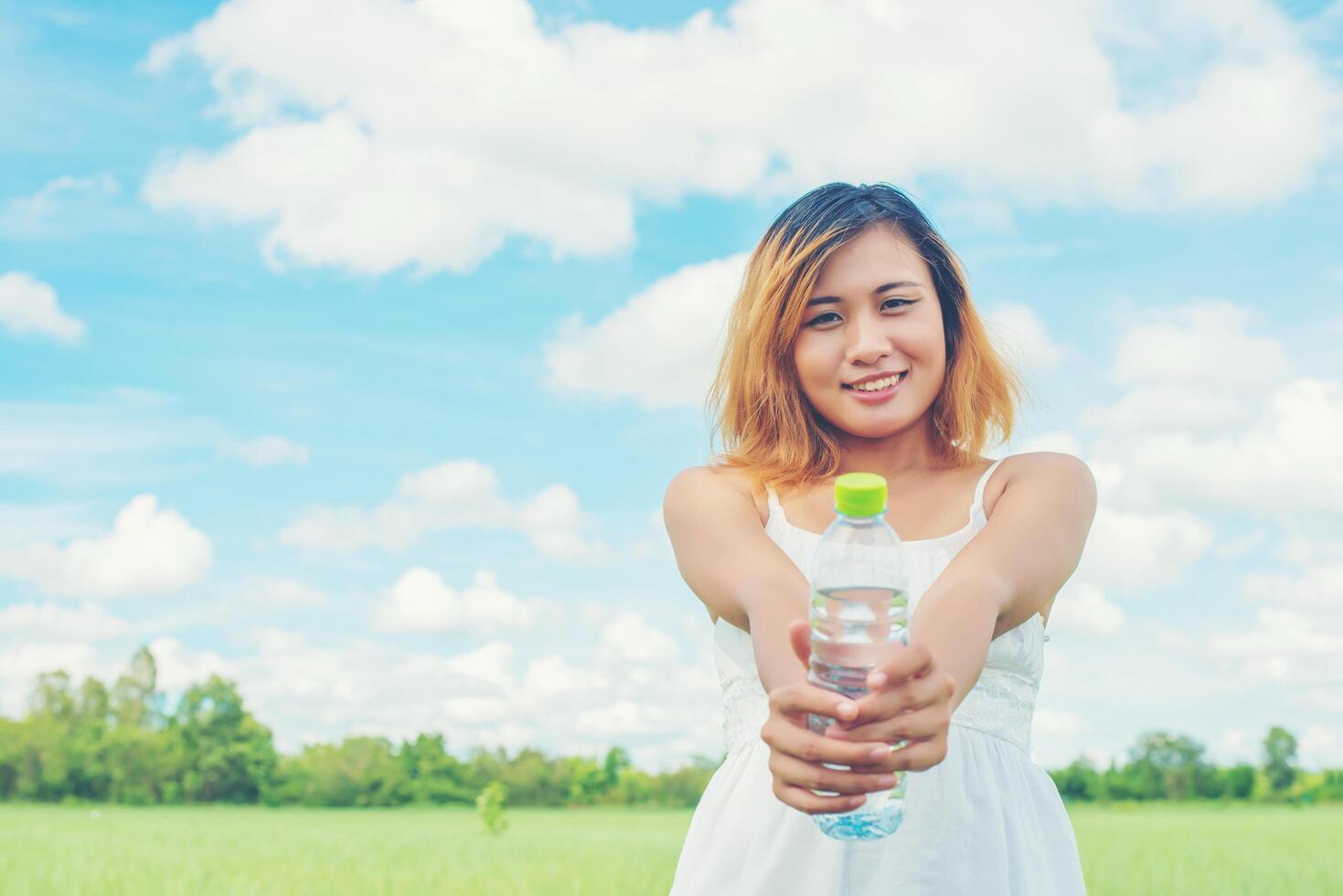 Frauen-Lifestyle-Konzept junge hübsche Frau, die weißes Kleid trägt, das eine Flasche Wasser auf Grünland-Smiley in die Kamera hält, sieht so frisch aus, genießt und ist glücklich. foto