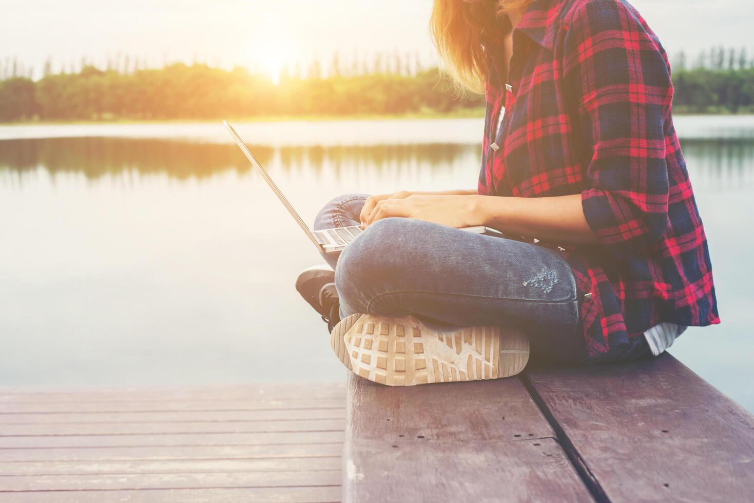 junge, glückliche hipster-frau, die mit ihrem laptop arbeitet, sitzt am pier und entspannt sich mit der natur. foto