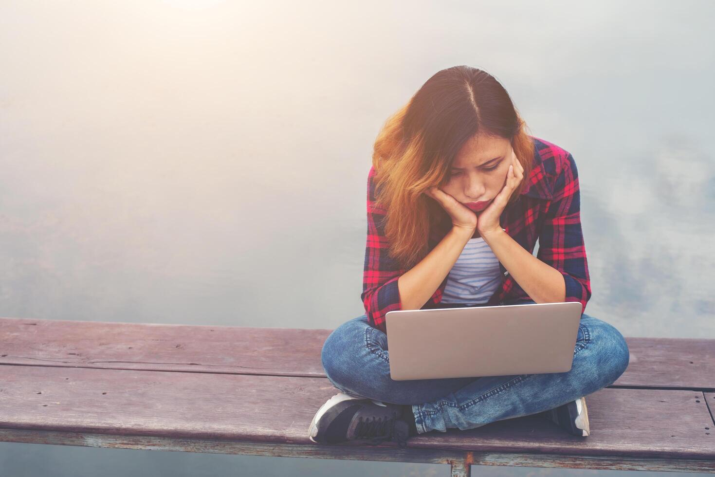 Junge Business-Hipster-Frau, die auf dem Pier sitzt, müde für die Arbeit, Blick auf den Laptop und langweilig. foto