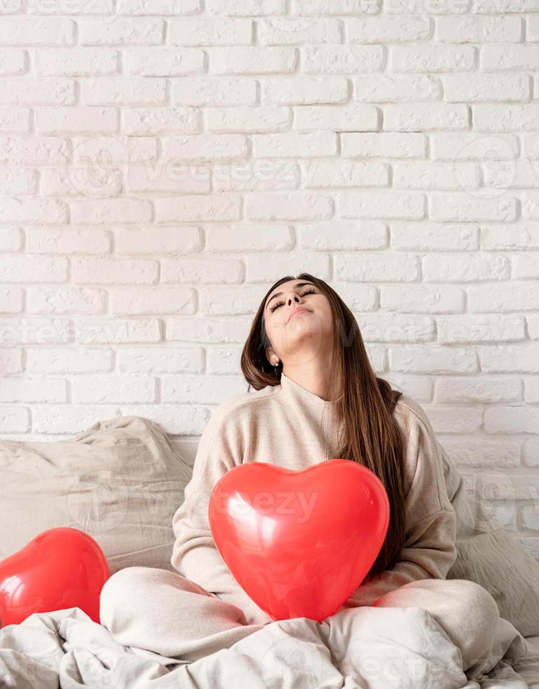 junge brünette frau sitzt im bett und feiert den valentinstag mit roten herzballons foto