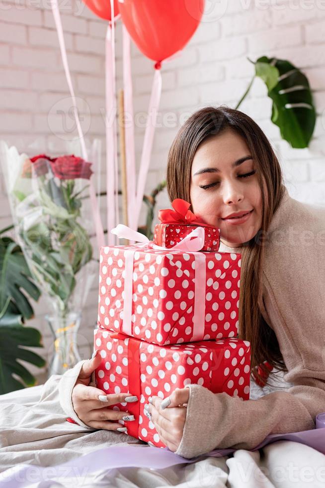 junge frau, die im bett sitzt und den valentinstag feiert und einen stapel von geschenken umarmt foto