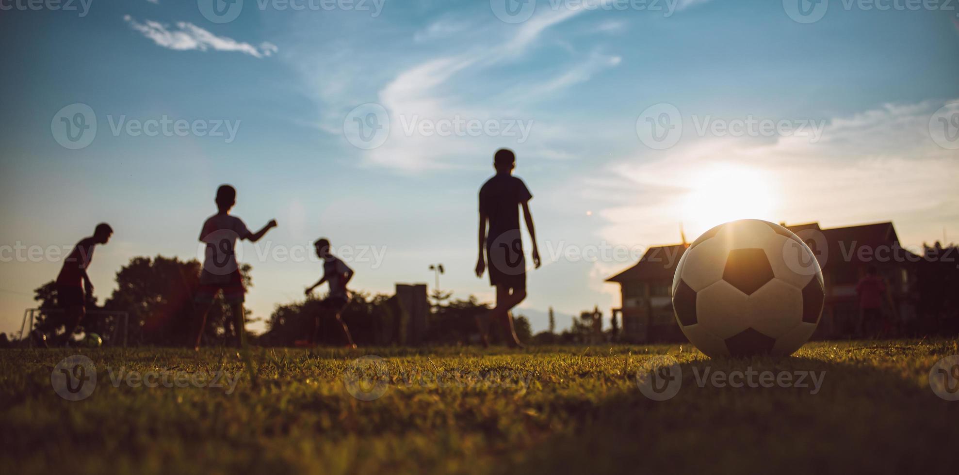 Silhouetten-Action-Sport im Freien von Kindern, die Spaß daran haben, Fußball zu spielen, um sich in der ländlichen Gegend der Gemeinde unter dem Dämmerungshimmel zu bewegen foto