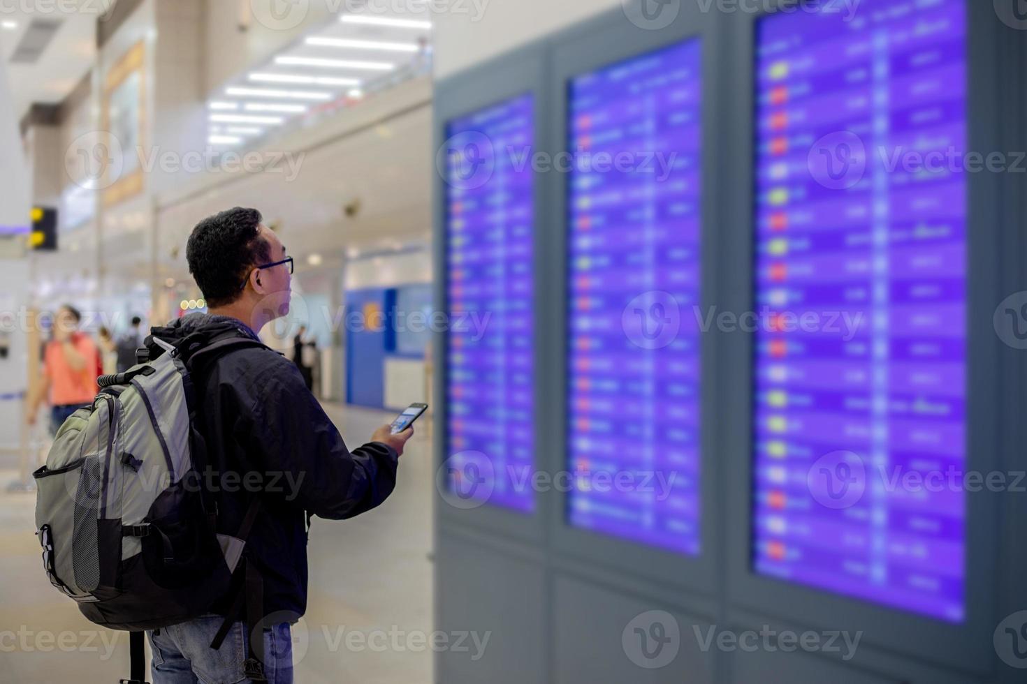 asiatischer mann mit rucksackreisendem, der das intelligente handy zum check-in am fluginformationsbildschirm in einem modernen flughafen verwendet foto