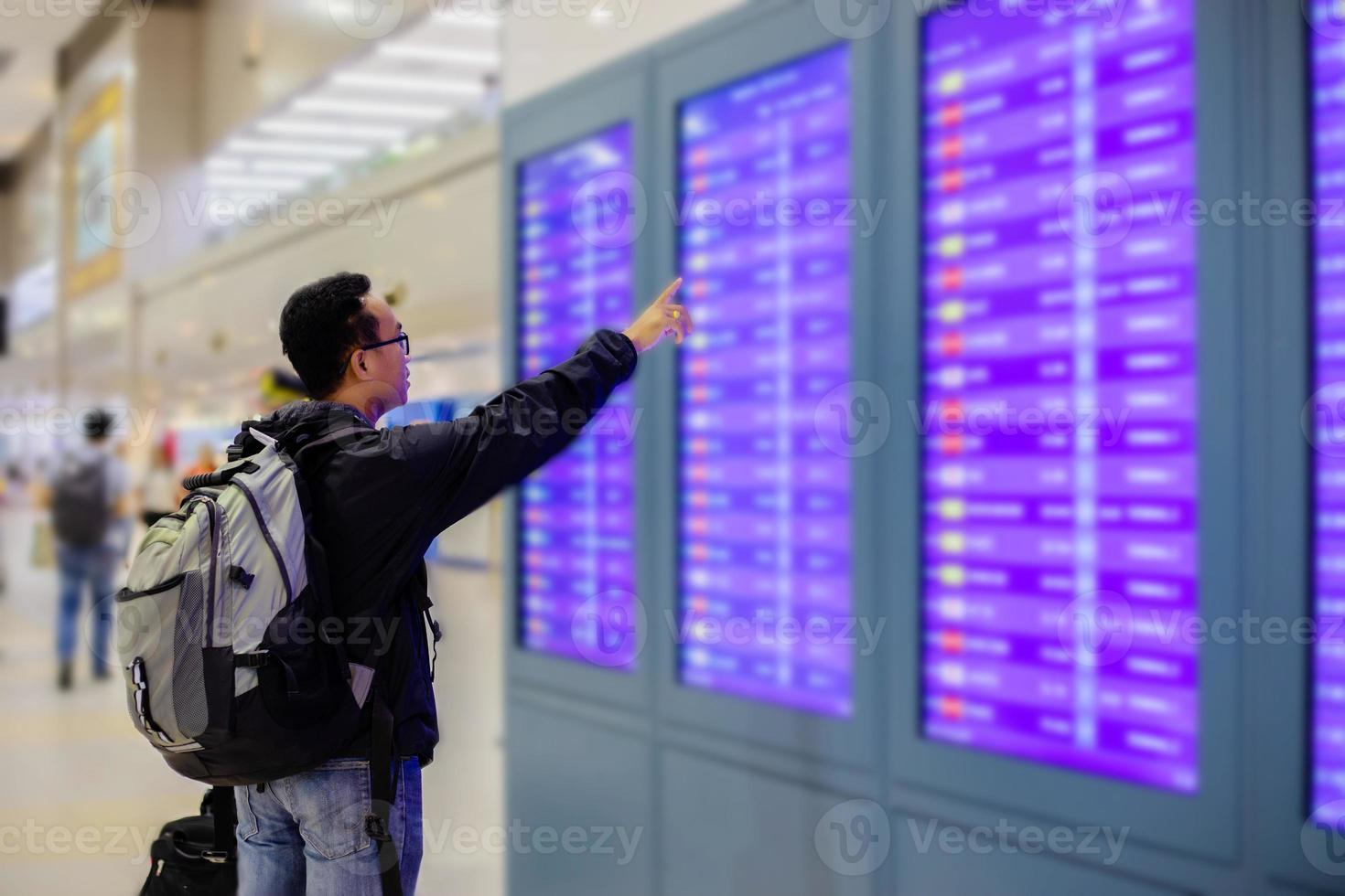 asiatischer mann mit rucksackreisendem, der das intelligente handy zum check-in am fluginformationsbildschirm in einem modernen flughafen verwendet foto