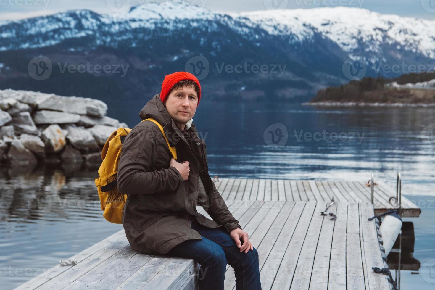 Mann mit einem gelben Rucksack, der einen roten Hut trägt, der auf einem Holzsteg auf dem Hintergrund von Berg und See sitzt foto