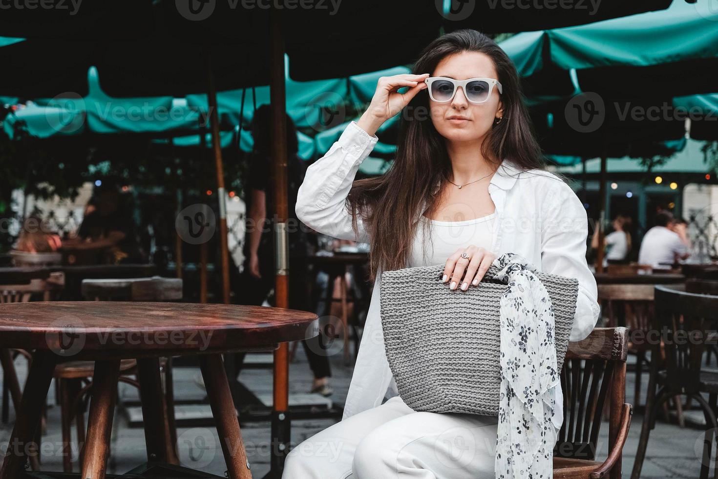 Frau mit Sonnenbrille und weißer Kleidung, die eine gestrickte Tasche hält, die am Tisch eines Straßencafés sitzt. kopieren, leerer platz für text foto