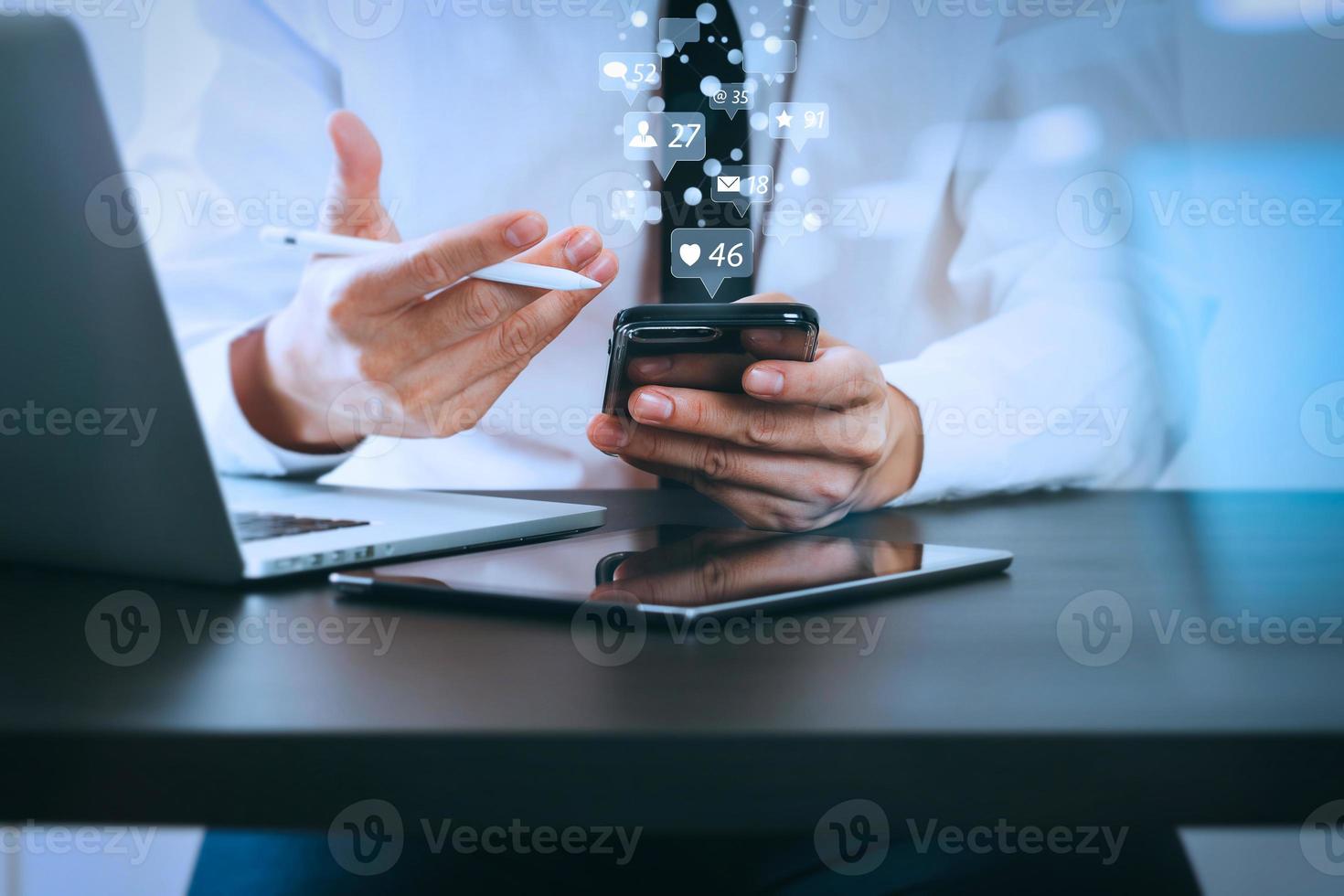 Nahaufnahme eines Geschäftsmannes, der mit einem Smartphone auf einem Holzschreibtisch in einem modernen Büro mit virtuellem Symboldiagramm mit reflektierter Glasansicht arbeitet foto