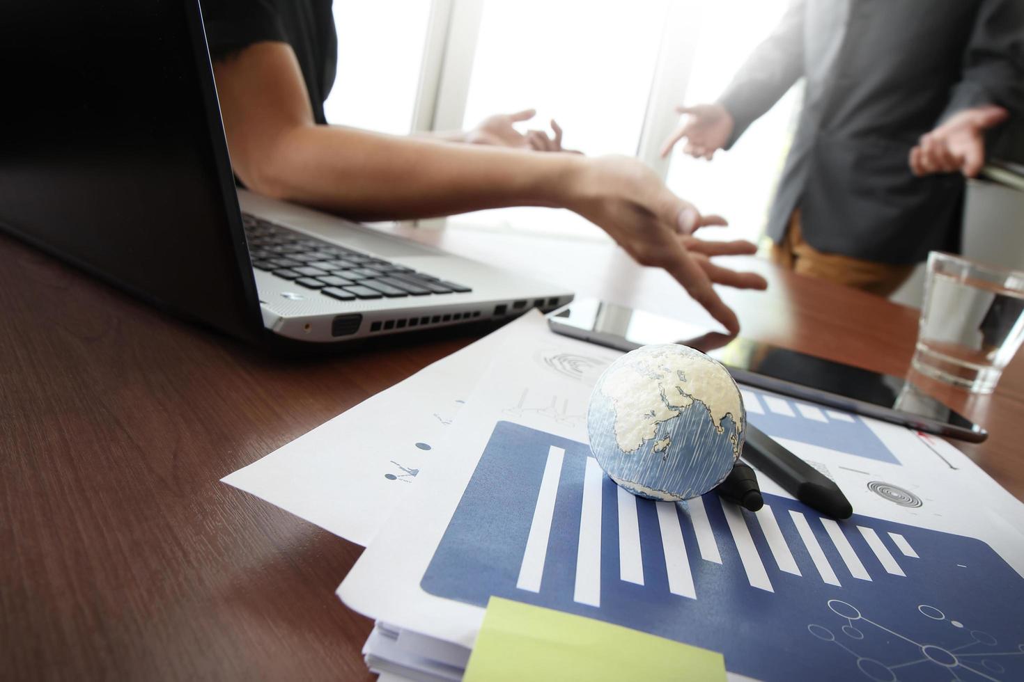 Geschäftsdokumente im Büro mit Textur der Welt und digitaler Tablet- und Laptop-Computer und zwei Kollegen, die Daten im Hintergrund diskutieren foto