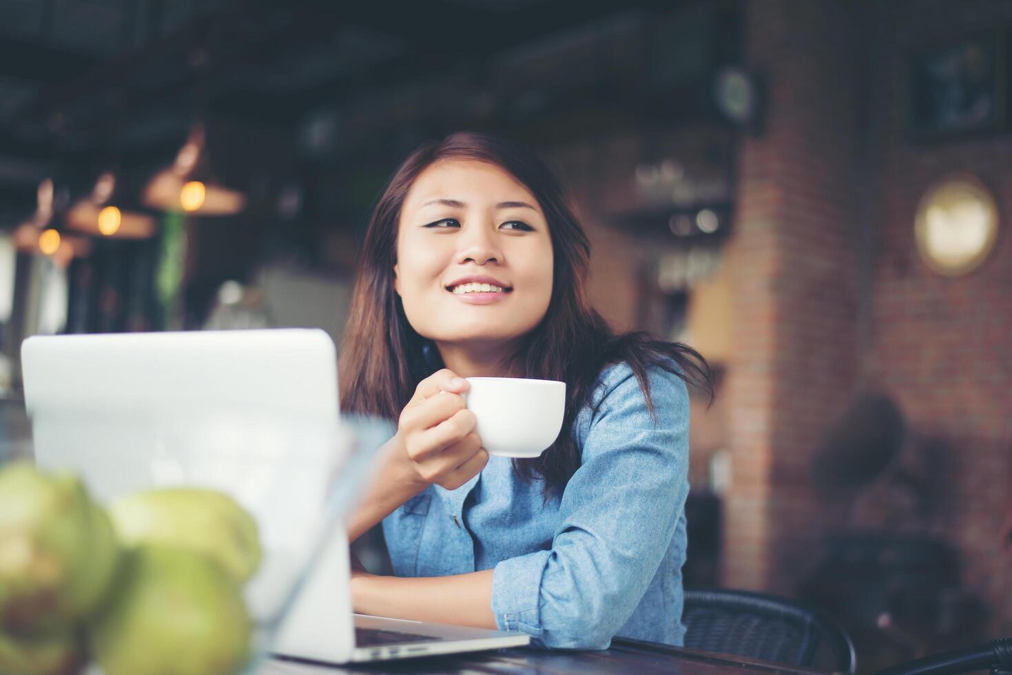 schöne junge hipster-frau, die in einem café sitzt und eine tasse kaffee hält, entspannt sich und spielt mit ihrem laptop, schaut weg, glücklich und macht spaß. Lifestyle-Konzept. foto