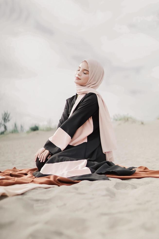 schönes islamisches weibliches modell, das hijab-mode trägt, ein modernes hochzeitskleid für muslimische frau, die im sand und am strand sitzt. Porträt eines asiatischen Mädchenmodells mit Hijab, das sich am Strand mit Bäumen amüsiert foto