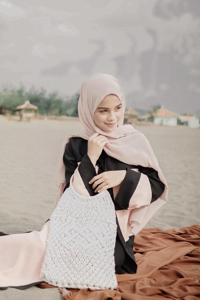 schönes islamisches weibliches modell, das hijab-mode trägt, ein modernes hochzeitskleid für muslimische frau, die im sand und am strand sitzt. Porträt eines asiatischen Mädchenmodells mit Hijab, das sich am Strand mit Bäumen amüsiert foto