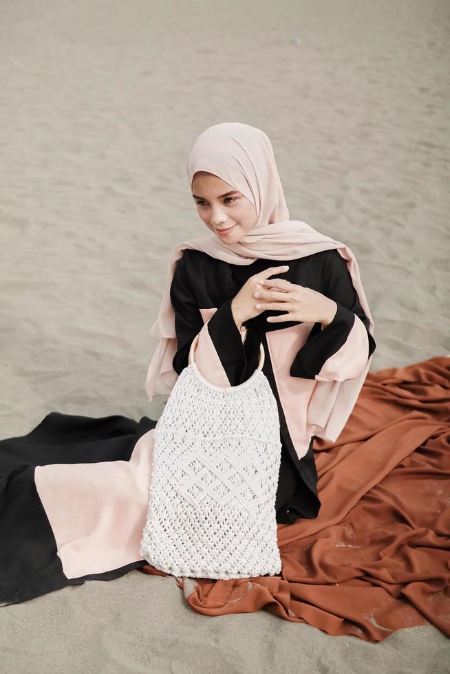 schönes islamisches weibliches modell, das hijab-mode trägt, ein modernes hochzeitskleid für muslimische frau, die im sand und am strand sitzt. Porträt eines asiatischen Mädchenmodells mit Hijab, das sich am Strand mit Bäumen amüsiert foto