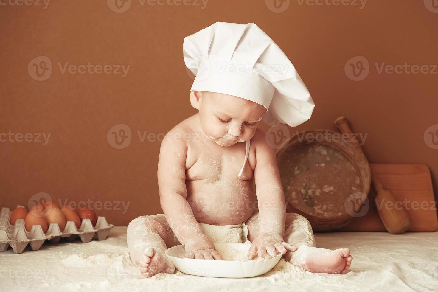 Kleiner Bäckerjunge mit Kochmütze sitzt auf dem Tisch und spielt mit Mehl auf braunem Hintergrund mit einem hölzernen Nudelholz, einem runden rustikalen Sieb und Eiern. kopieren, leerer platz für text foto