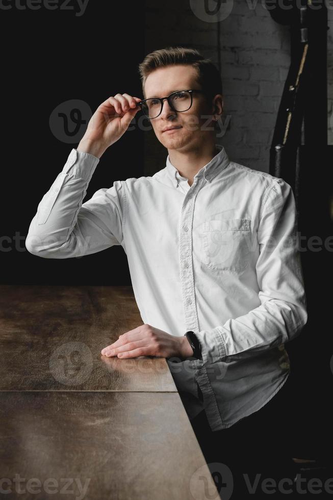 Junger Mann mit Brille und weißem Hemd, der auf dunklem Hintergrund am Tisch sitzt. kopieren, leerer platz für text foto