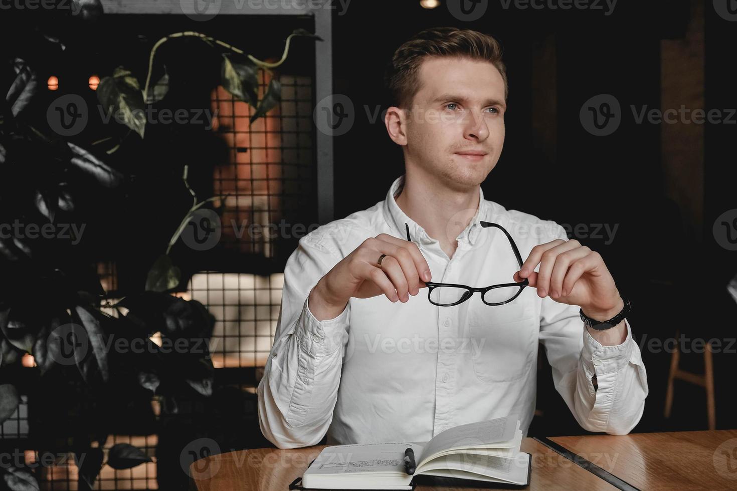 Junger Mann mit weißem Hemd und Brille, der am Tisch sitzt und einen Stift hält, macht sich auf dunklem Hintergrund im Büro Notizen zu seiner Agenda. kopieren, leerer platz für text foto