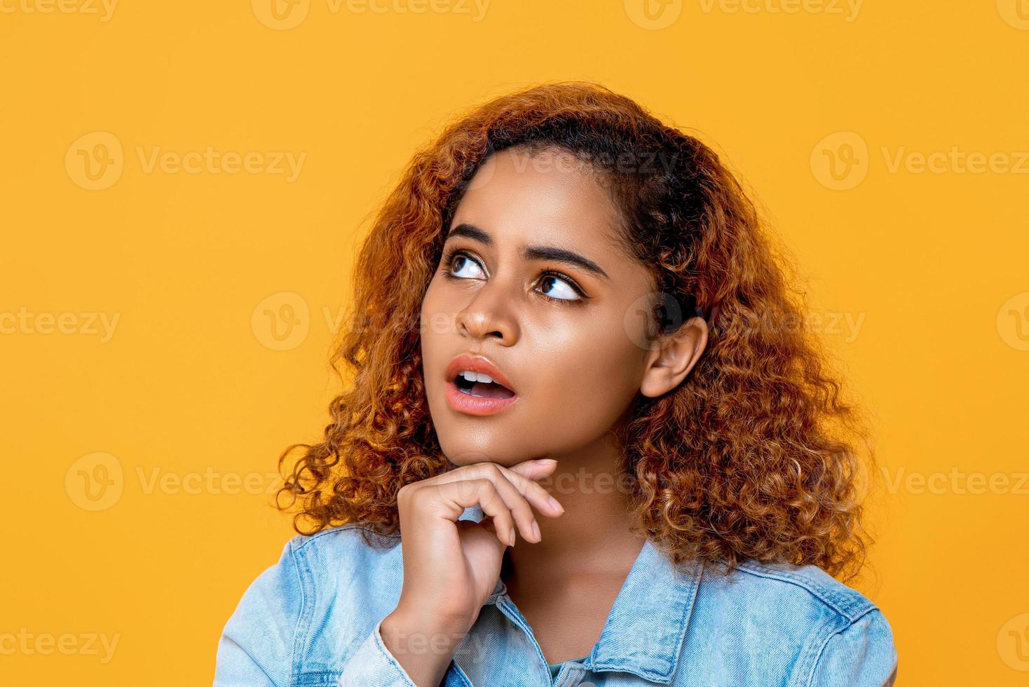 gemischte afrikanische frau, die mit der hand am kinn und den augen nach oben schaut, die isoliert auf gelbem hintergrund in den raum schauen foto