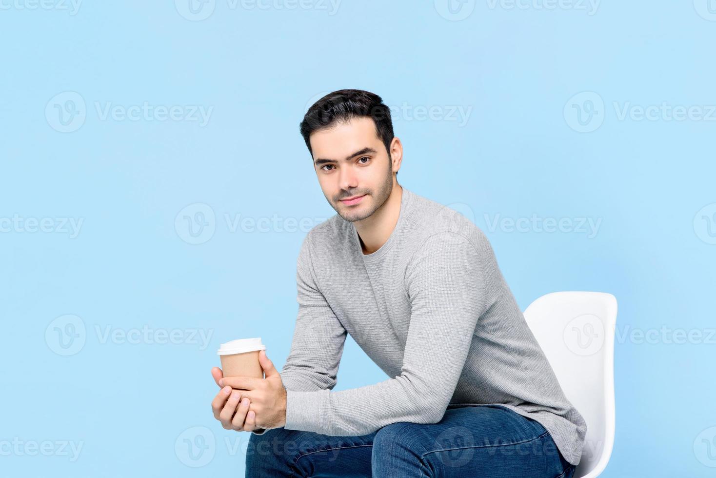 Porträt eines jungen gutaussehenden kaukasischen Mannes, der in die Kamera blickt, während er sitzt und eine Kaffeetasse isoliert auf hellblauem Studiohintergrund hält foto