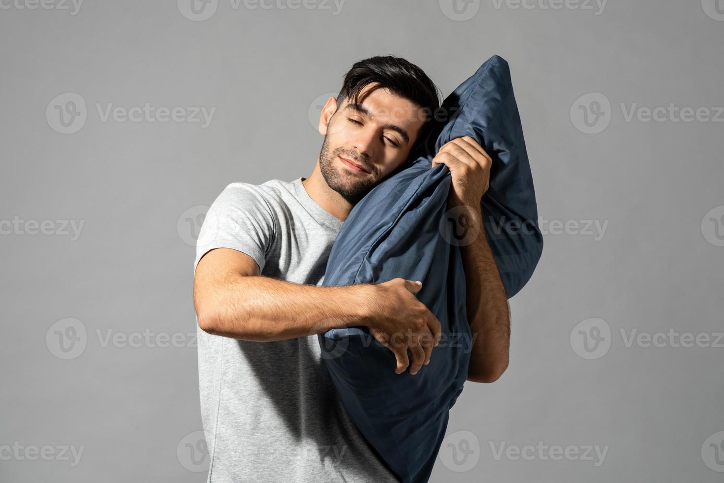 Isoliertes Porträt eines lächelnden jungen Mannes, der ein Kissen hält, das seine Augen schließt und auf hellgrauem Studiohintergrund träumt foto