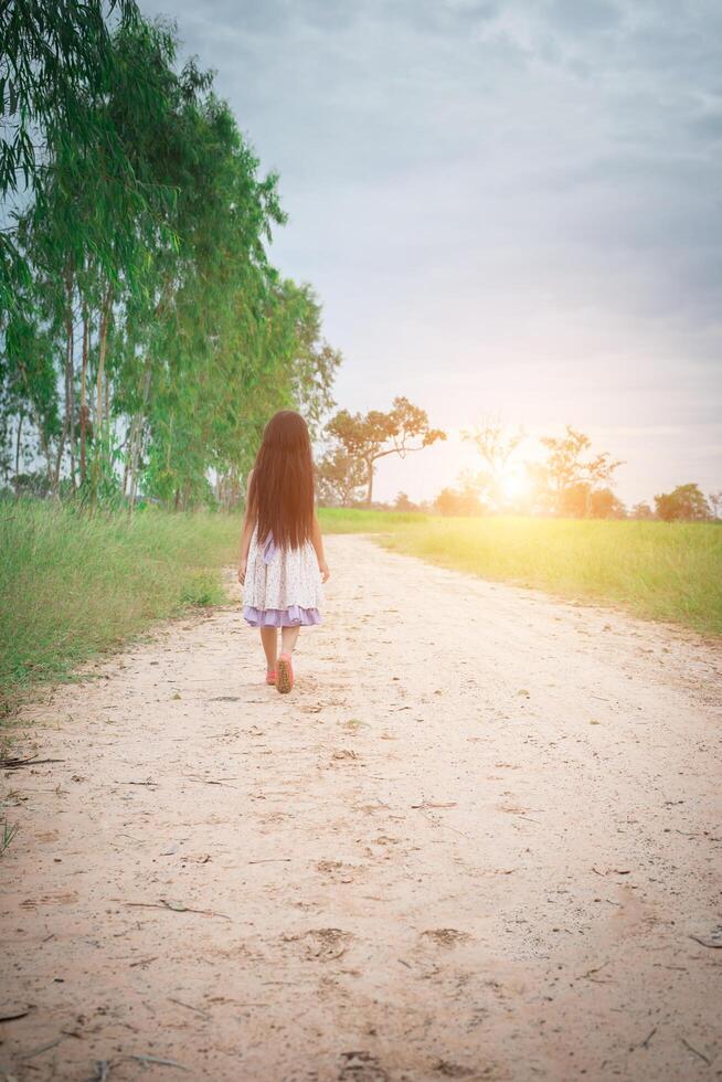 Kleines Mädchen mit langen Haaren, das Kleid trägt, geht von Ihnen weg auf der Landstraße. foto