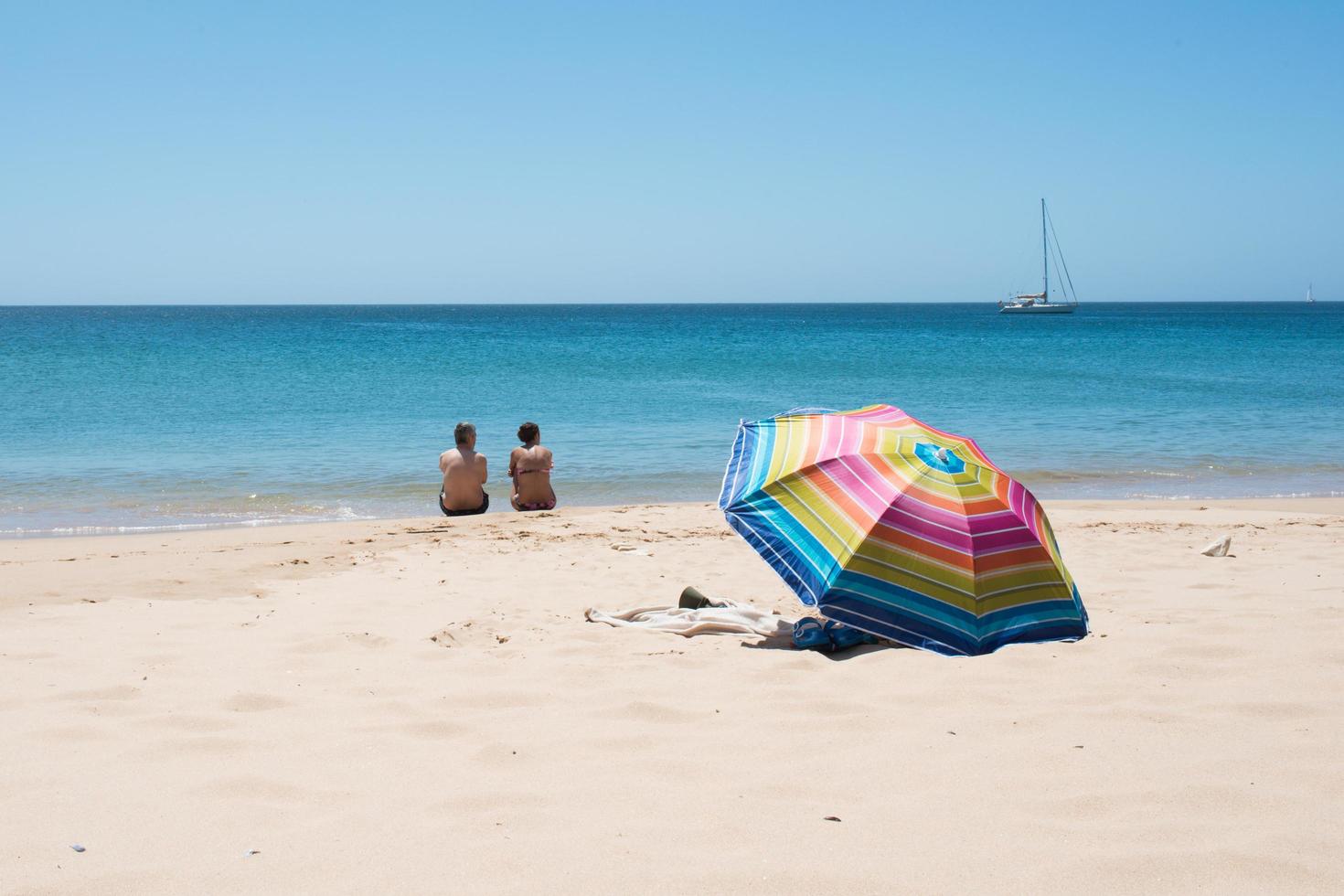 erwachsenes paar sitzt an einem strand mit blick auf das meer. von hinten gesehen, in der nähe ein bunter sonnenschirm, menschenleer, in der ferne eine yacht. Strand von Sagres, Algarve, Portugal. foto