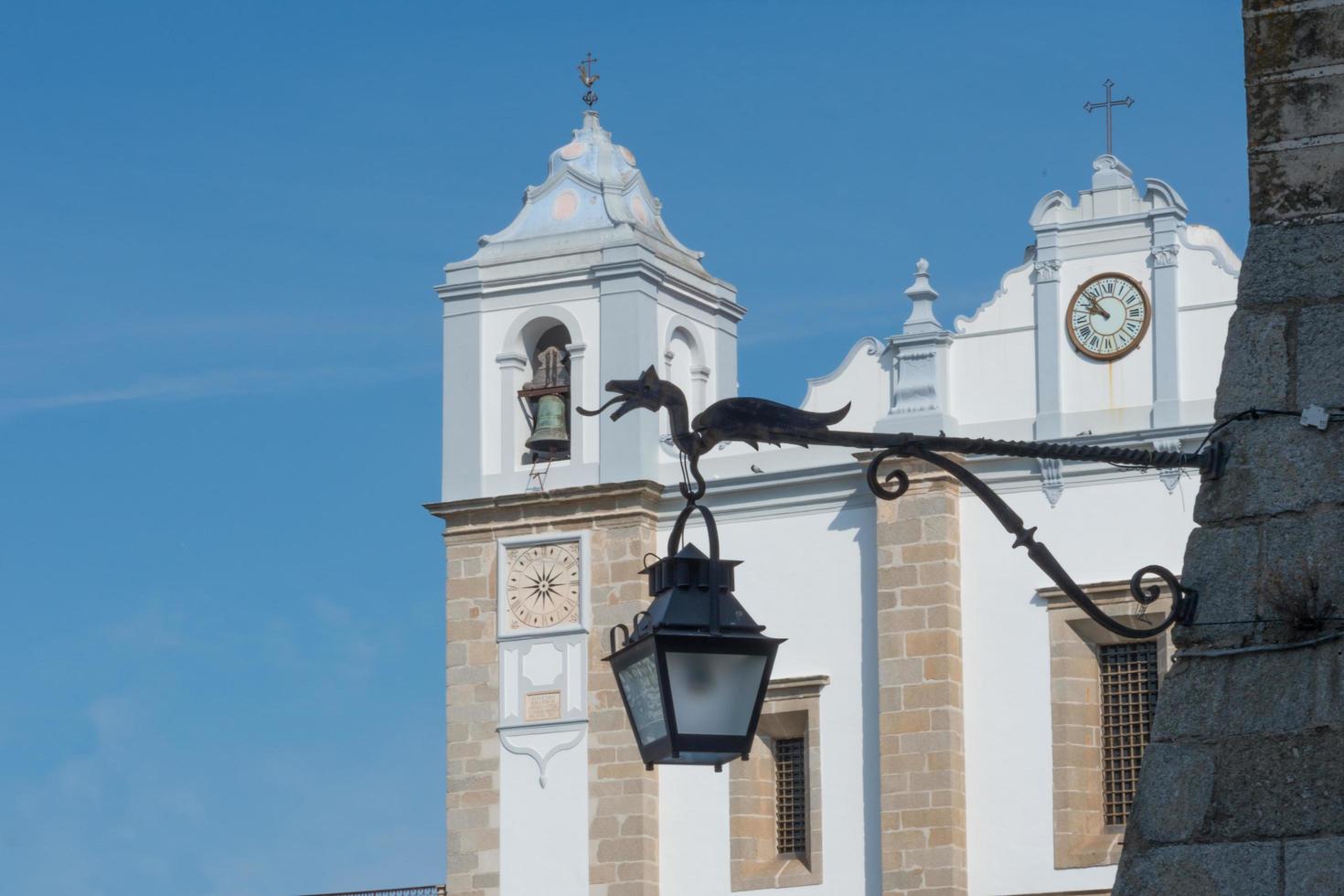 Nahaufnahme einer Straßenlaterne mit einer Drachenskulptur. im hintergrund kirche am giraldo-platz, evora, portugal. blauer Himmel foto