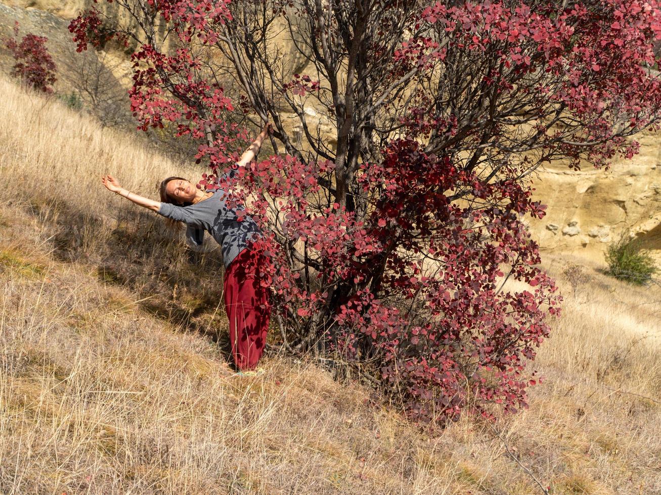 Lächelnde Frau mit roter indischer Hose in der Nähe eines Baumes mit roten Blättern praktiziert Yoga, Herbstporträt foto