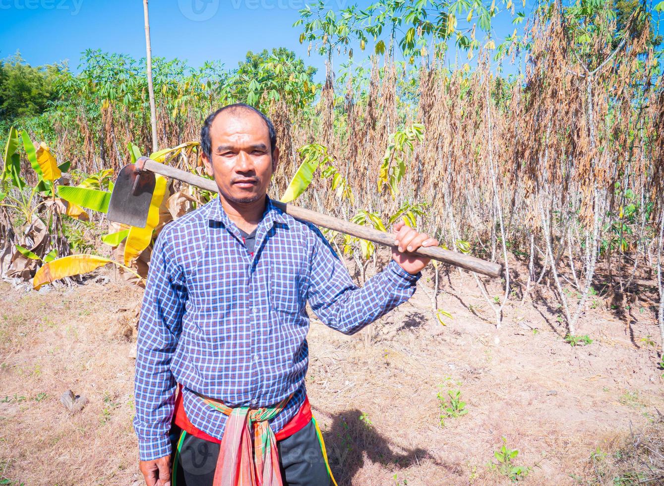 junger asiatischer mann, bauer, voll bekleidet. um die Sonne auszublenden, eine Machete zu halten, in die Kamera zu schauen und leicht zu lächeln. junger Mann, der auf dem Bauernhof steht foto