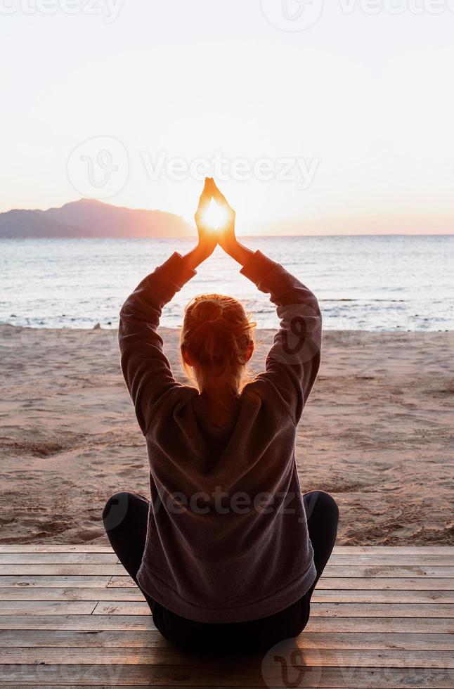 Junge gesunde Frau, die Yoga praktiziert und Händchen hält, in Meditationspose mit Sonne zwischen ihnen am Strand bei Sonnenaufgang foto