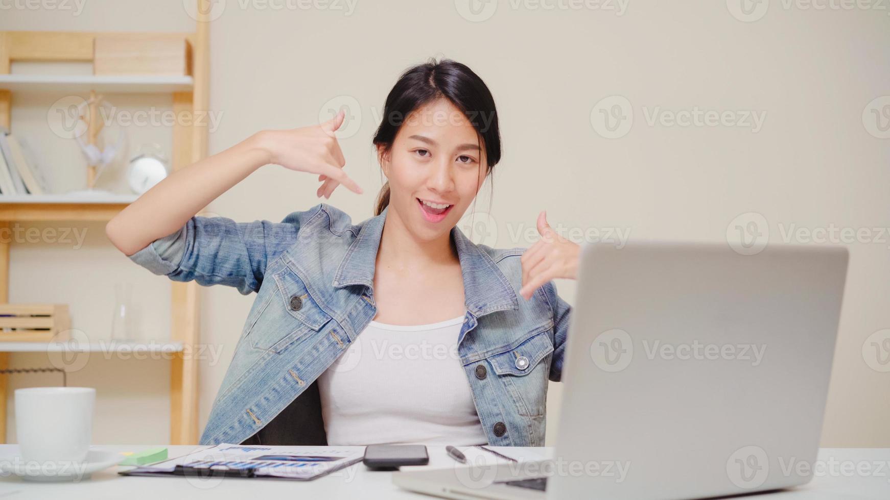 junge asiatische frau, die zu hause mit laptop auf schreibtisch im wohnzimmer arbeitet. asien geschäftsfrau erfolgsfeier gefühl glücklich tanzen im büro zu hause. zeit zu hause genießen. foto