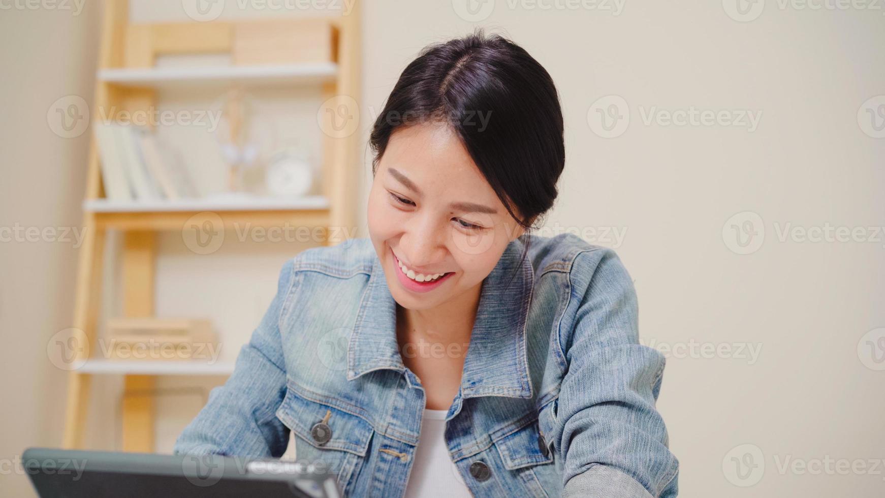 junge asiatische frau, die mit einem tablet arbeitet und soziale medien überprüft, während sie sich zu hause auf dem schreibtisch im wohnzimmer entspannt. zeit zu hause genießen. foto