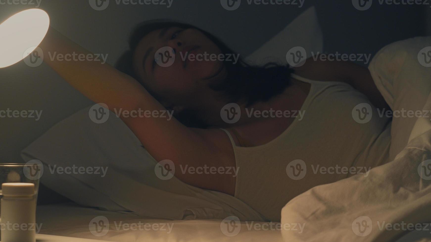 schöne asiatische junge frau, die gemütlich in ihrem schlafzimmer schläft. indische weibliche hand, die den lichtschalter in der nähe des bettes im zimmer zu hause in der späten nacht zum schlafen bereit einschaltet. süße träume und entspannungskonzept. foto