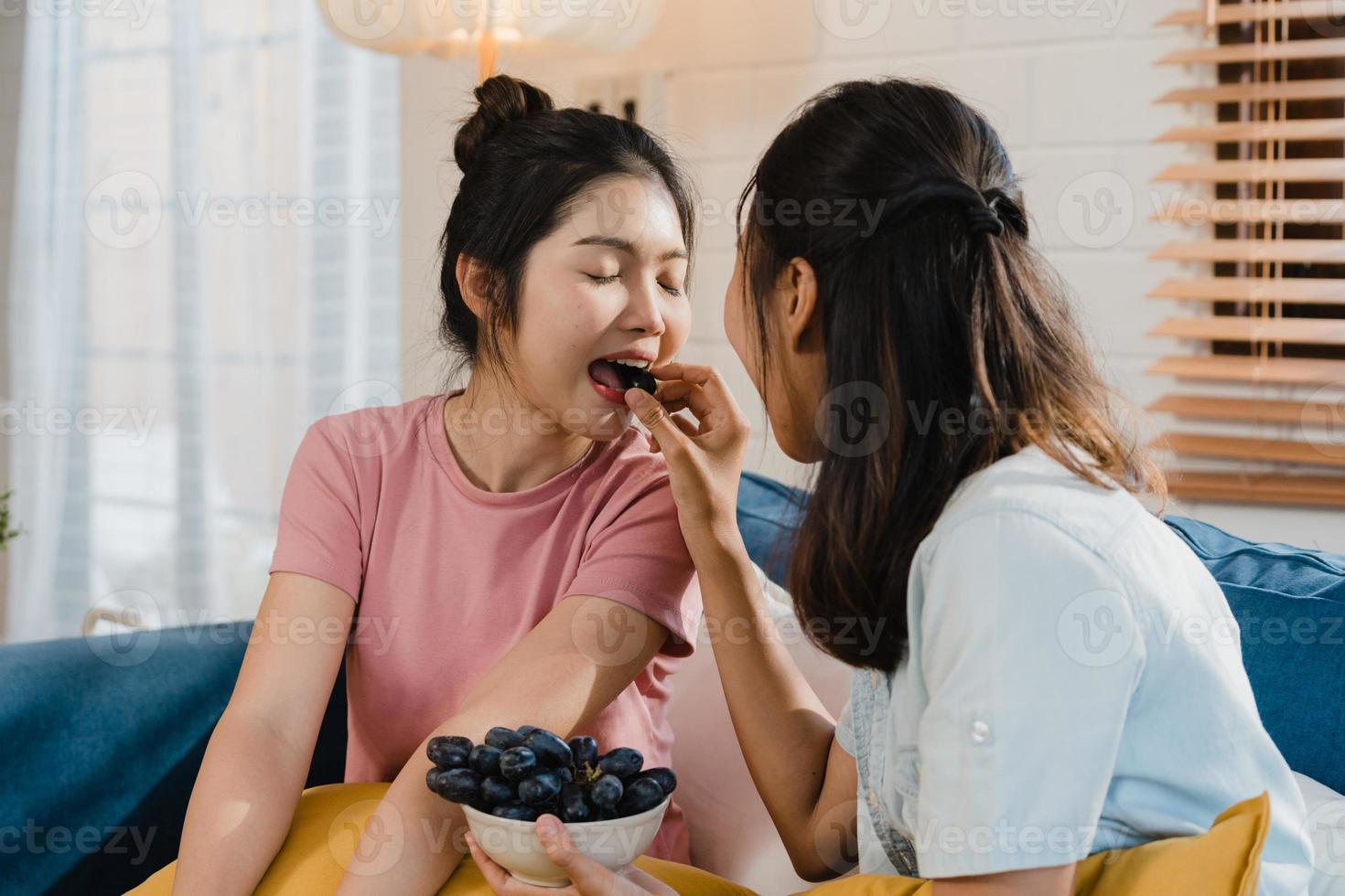 asiatische lesbische lgbtq-frauenpaare essen gesundes essen zu hause, junge asiatische liebhaberinnen fühlen sich glücklich, trauben zusammen zu füttern, während sie morgens zu hause sofa im wohnzimmer liegen. foto