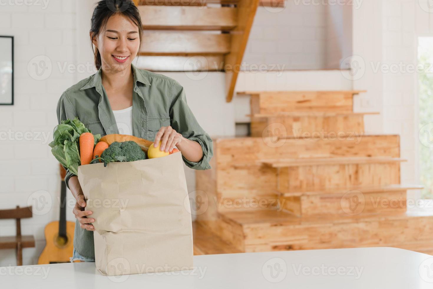 asiatische frauen, die zu hause einkaufspapiertüten halten, junge asiatische mädchen kaufen gerne gemüse und obst, gesundes und organisches produkt aus dem supermarkt, legen es morgens in die küche. foto
