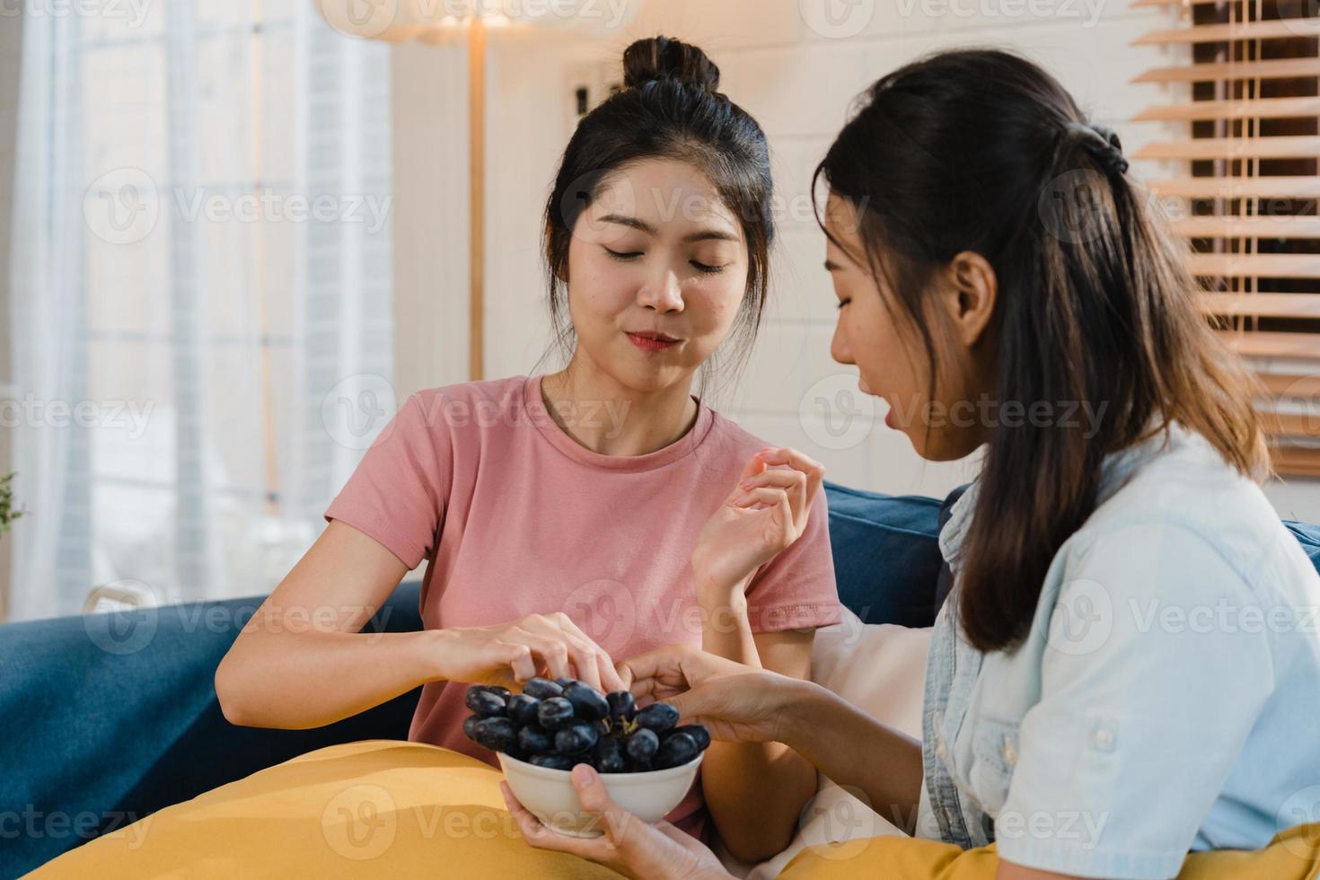 asiatische lesbische lgbtq-frauenpaare essen gesundes essen zu hause, junge asiatische liebhaberinnen fühlen sich glücklich, trauben zusammen zu füttern, während sie morgens zu hause sofa im wohnzimmer liegen. foto