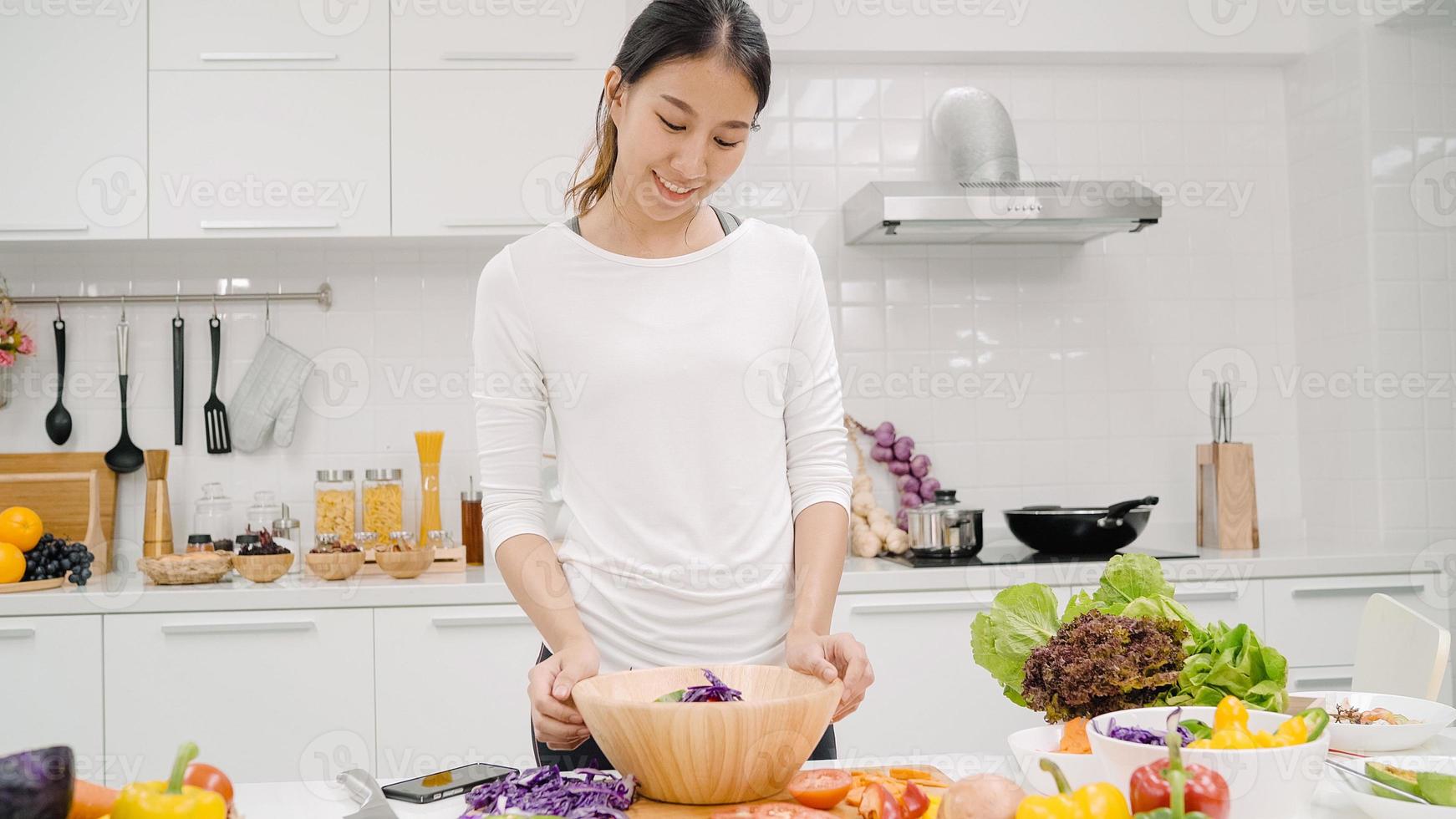 junge asiatische frau, die salat gesundes essen in der küche macht, schöne frau im gelegentlichen gebrauch bio-gemüse viel ernährung bereitet salat für fitten körper zu hause zu. gesundes lebensmittelkonzept. foto