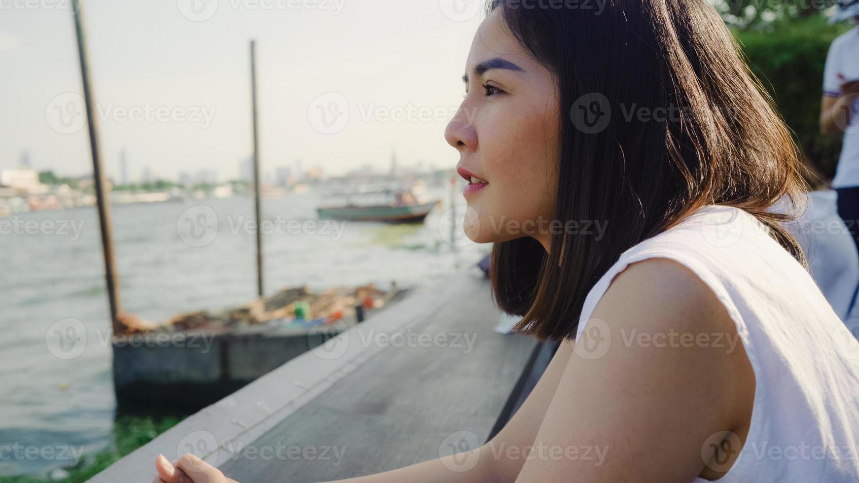 junge asiatische bloggertouristin mit lässigem blick auf die vorderseite des hafens und des bootes auf dem fluss fühlen sich entspannend, glücklich, freudig reisen im café in der stadt, lebensstil touristenreise urlaubskonzept. foto