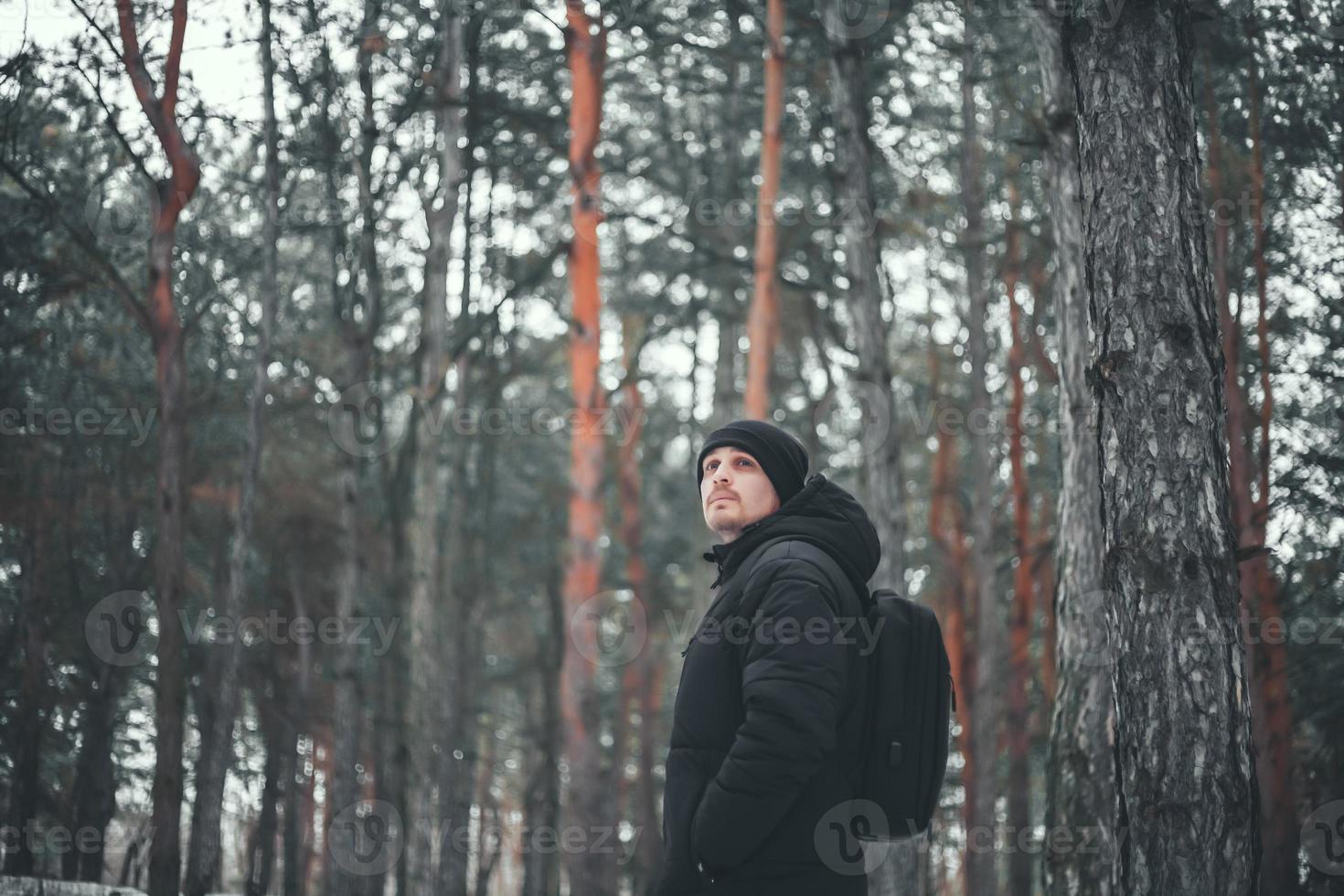 Ein junger attraktiver Typ mit kurzen Haaren in einem schwarzen Wintermantel posiert vor dem Hintergrund eines Winterwaldes. foto