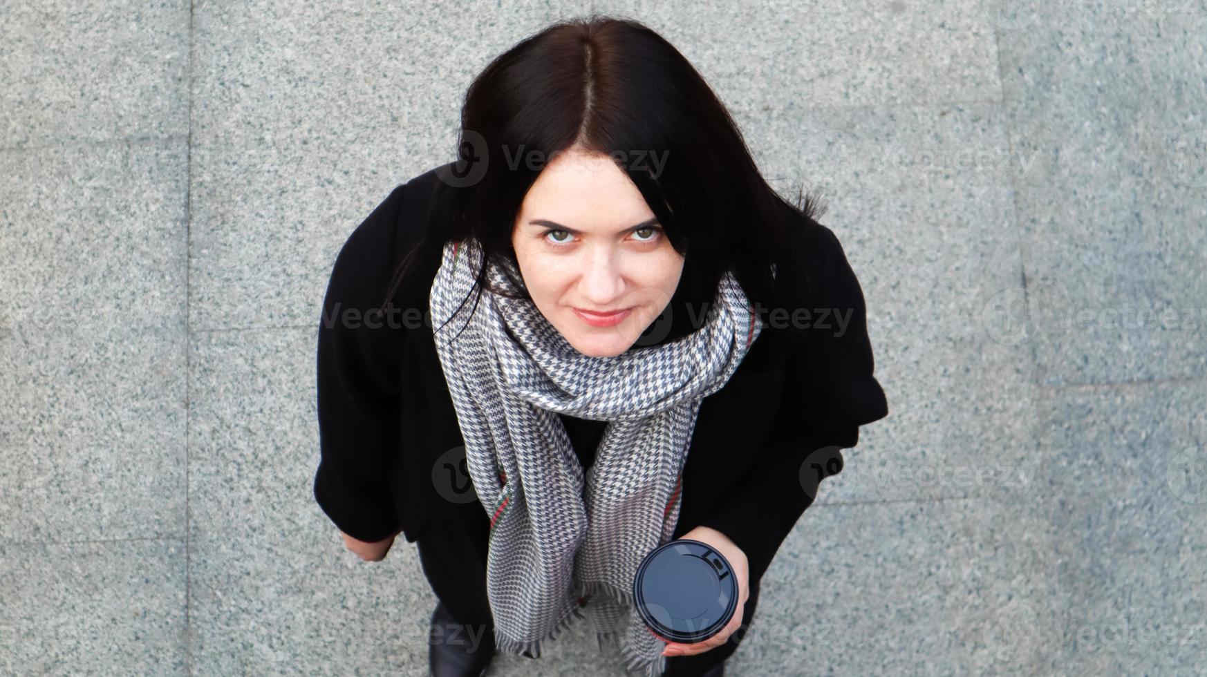 Junge stilvolle Frau in Mantel und Schal trinkt morgens heißen Kaffee in einem roten Öko-Papierglas im Freien in einem Herbstpark. Porträt einer jungen Frau mit einer Kaffeetasse zum Mitnehmen, Draufsicht. foto