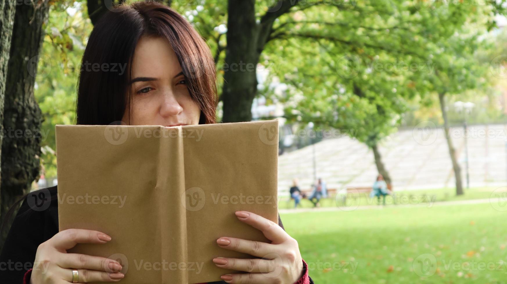 Porträt einer jungen Frau, die ein aufgeschlagenes Buch ohne Titel vor ihr Gesicht hält. Brünette, die an einem sonnigen Tag im Park die Hälfte ihres Gesichts mit einem Buch bedeckt. bildungs- und menschenkonzept, selektiver fokus. foto