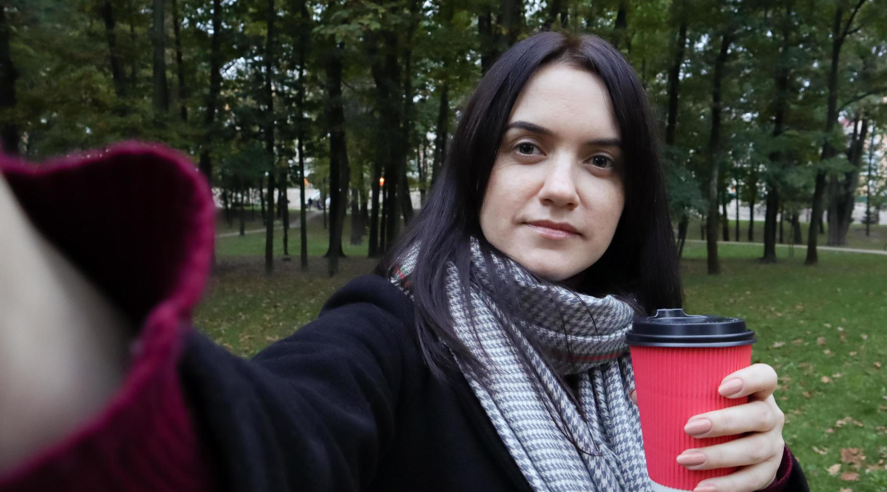 Schöne junge kaukasische Frau in einem Mantel mit einem Pappbecher Kaffee zum Mitnehmen, der ein Selfie macht oder Fotos von sich selbst für einen Blog mit einem Smartphone im Freien in einem Herbstpark macht.