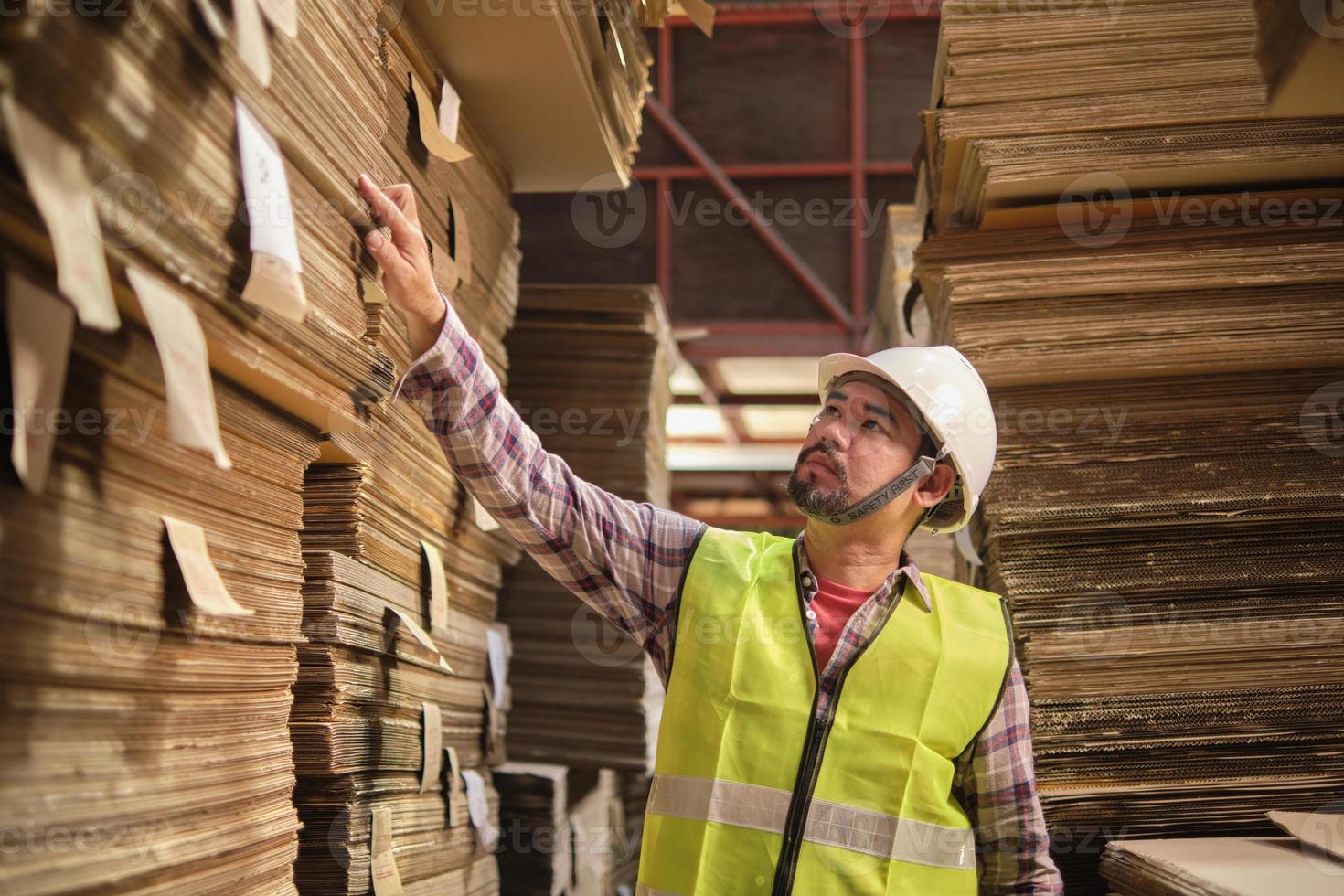 asiatischer männlicher arbeiter in sicherheitsuniform und schutzhelm, vorgesetzter inspiziert die verpackungslagerbestellung für die logistik im werkslager, stapel von stapelpapierherstellung, recyclingproduktionsindustrie. foto