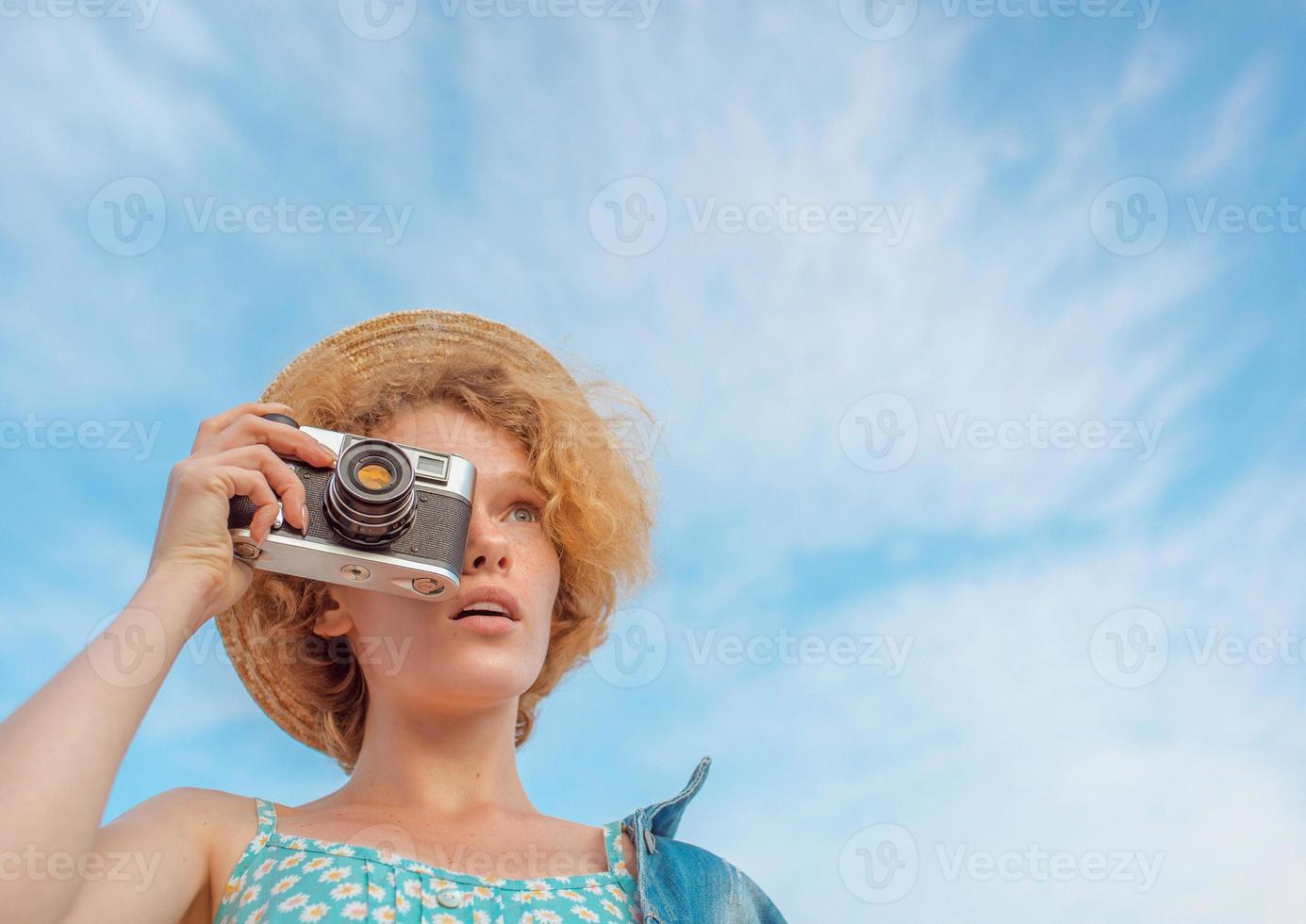 junge lockige rothaarige Frau mit Strohhut, blauem Sommerkleid und Jeansjacke, die mit einer Vintage-Kamera steht und Fotos auf blauem Himmelshintergrund macht. spaß, sommer, mode, schießen, reisen, jugendkonzept