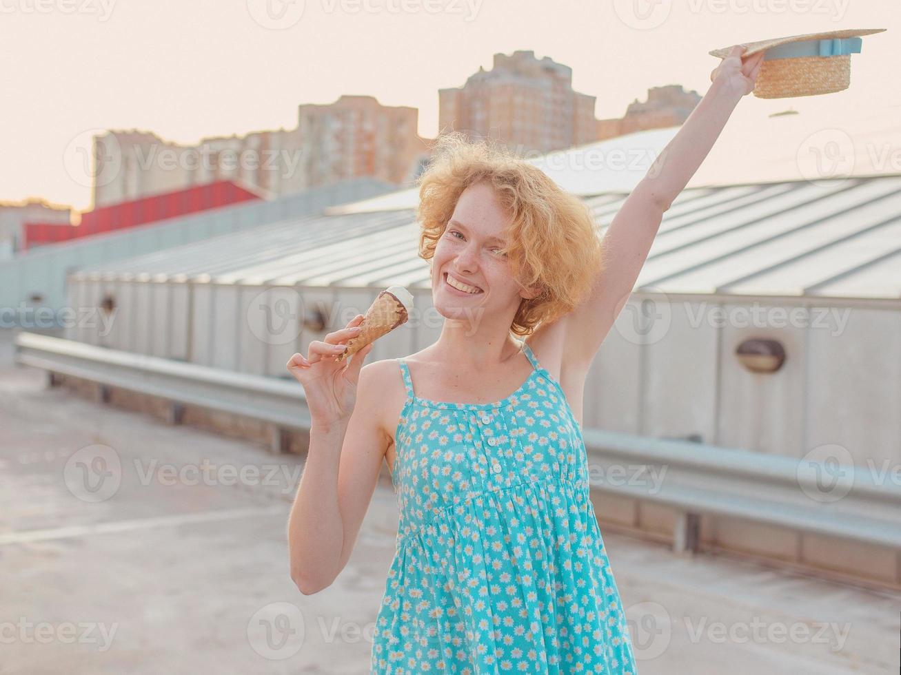 junge glückliche, fröhliche, lockige rothaarige frau mit strohhut, blaues sommerkleid, das eis auf wolkenkratzerdach isst. spaß, lebensstil, urban, modern, dach, stadt, sommer, mode, jugendkonzept foto