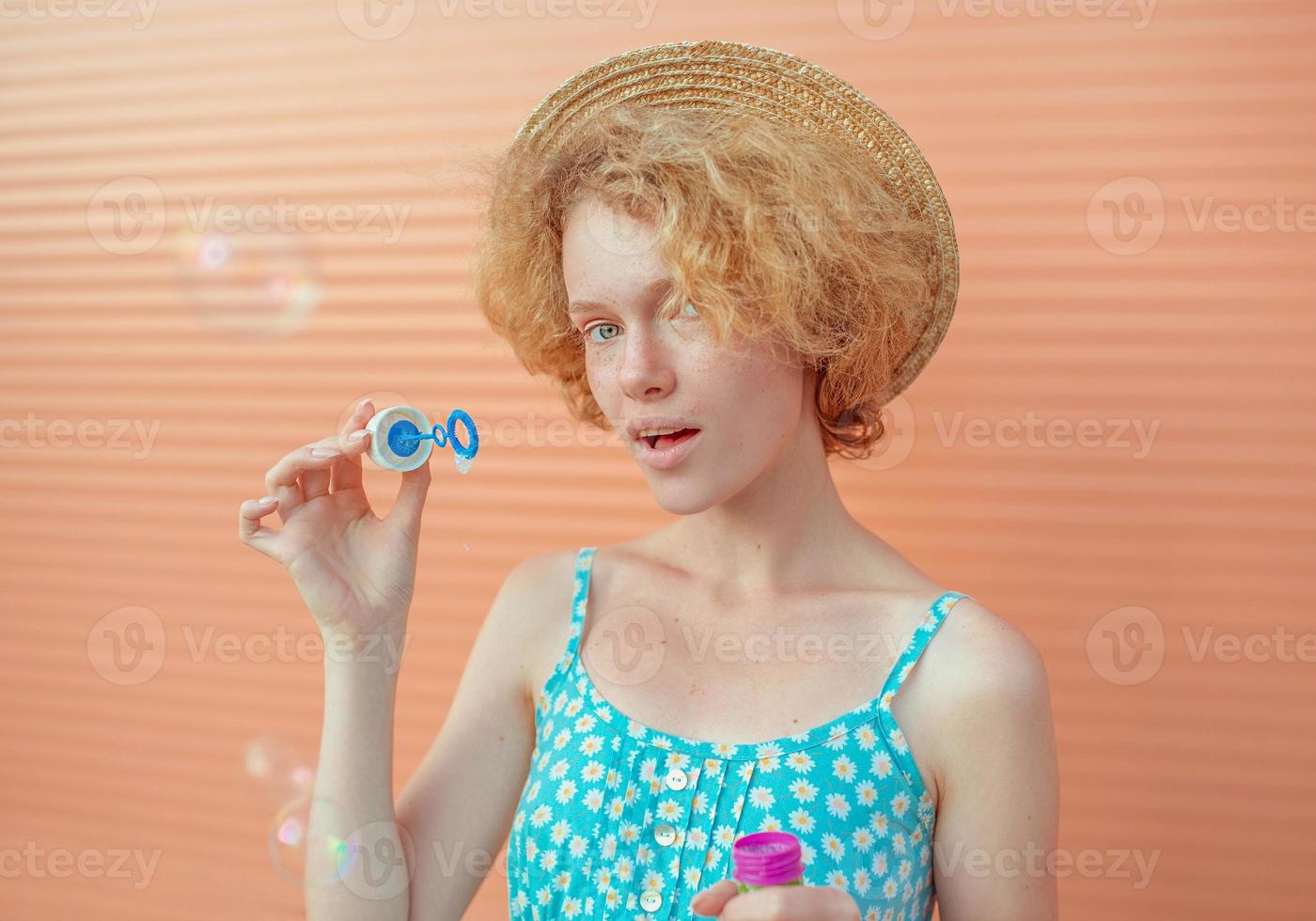 junge, fröhliche, lockige rothaarige frau in blauem sommerkleid und strohhut mit blasen auf beigem hintergrund. spaß, sommer, mode, jugendkonzept foto