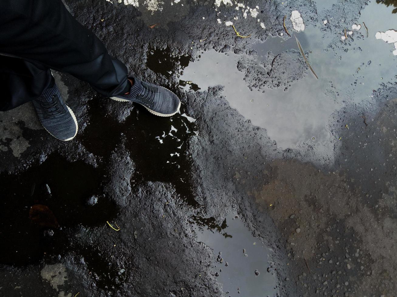 ein Foto von den Füßen einer Person, die Trainingshosen und -schuhe auf dem Asphalt auf einer mit Schlaglöchern überfluteten und beschädigten Straße trägt