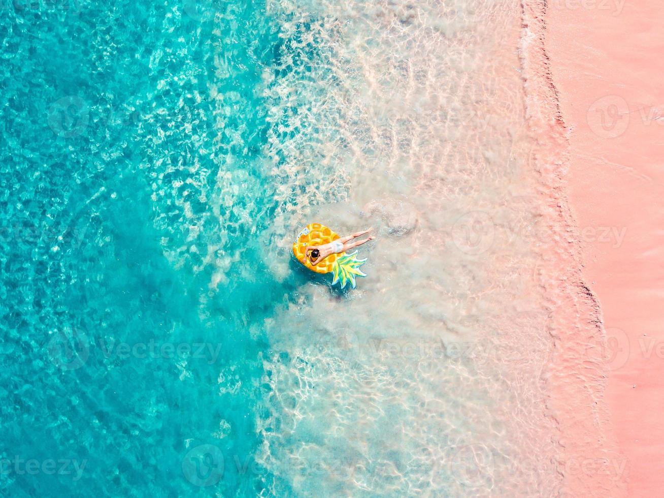 Drohnen-Luftaufnahme eines Mädchens, das auf einer aufblasbaren Ananas am exotischen tropischen rosafarbenen Strand schwimmt foto