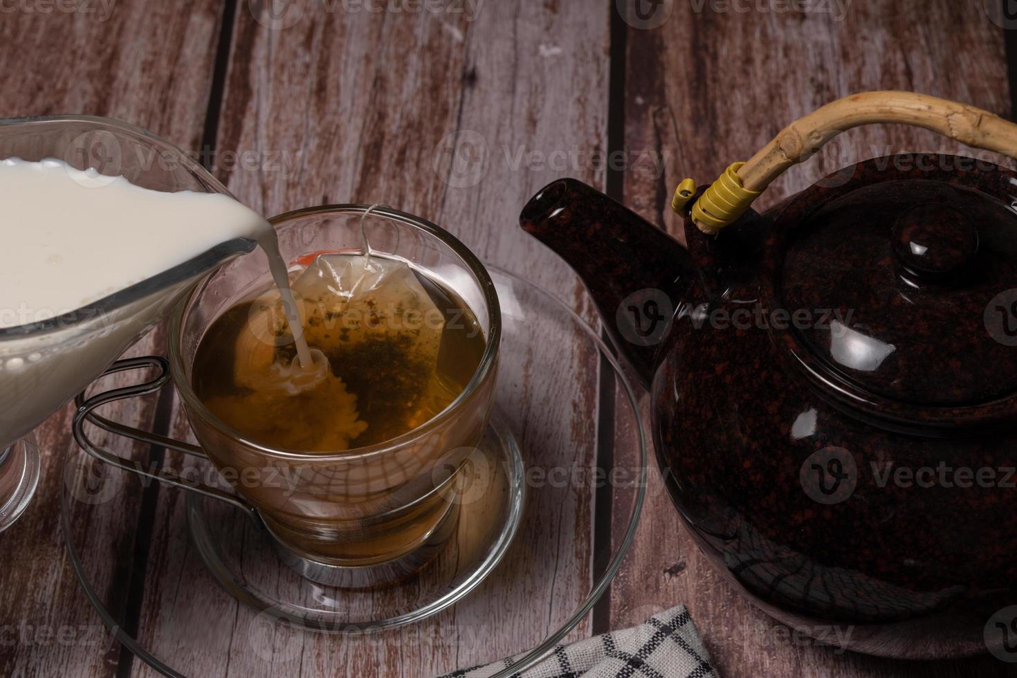 Teetasse und Teekanne, die eine Wolke heißer Milch auf einen Holztisch gießen foto