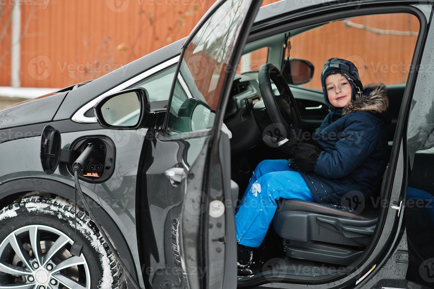 kleiner junge sitzt im auto und lädt elektrofahrzeug im hof des hauses im winter auf. foto