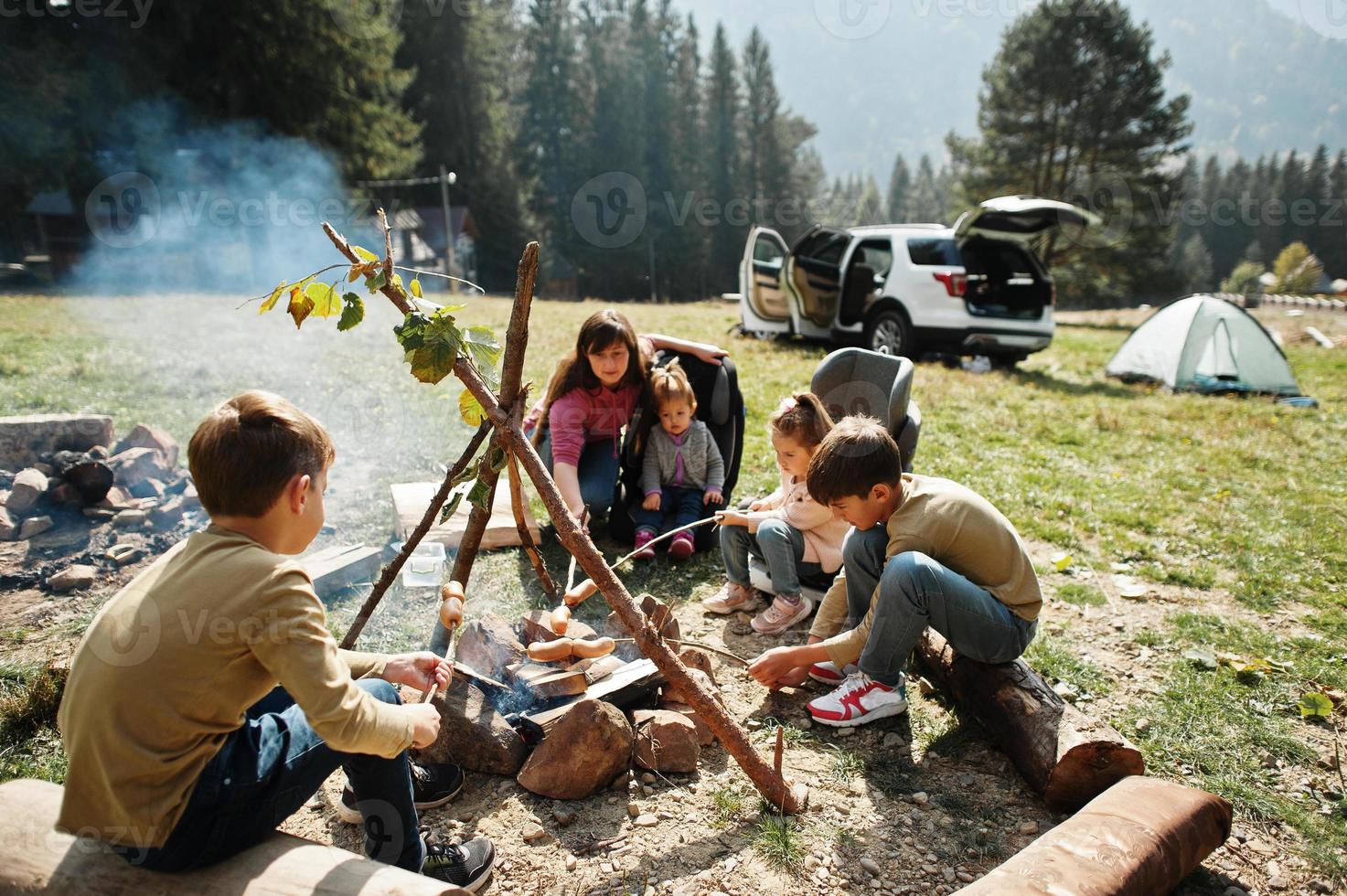 Familienfeuer im Berg. Bratwürste. Mutter mit vier Kindern Camping. Herbstwanderung und Lagerwetter. Erwärmen und Kochen in der Nähe einer Flamme zusammen. foto